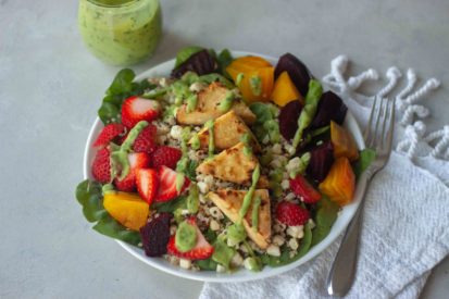 Sweet Corn, Berry, and Roasted Beet Quinoa Spinach Salad a plate of spinach, quinoa, sweet corn, tofu, roasted beets, sliced strawberries and avocado cilantro dressing drizzled and in jar.