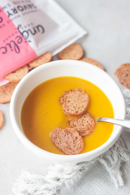 Bowl of Golden Turmeric Butternut Squash Soup topped with Angelic Bakehouse Sprouted Pickled Sweet Onion & Beet Crisps