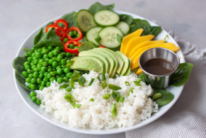 Plate with spinach, rice, sweet peas, red peppers, sliced cucumber, avocado, mango, and a cup of Asian dressing.