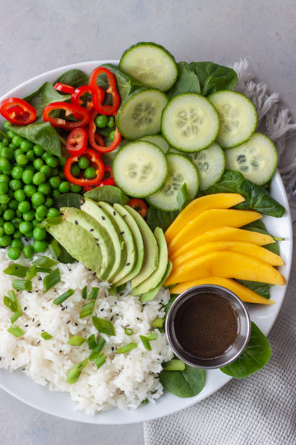 Plate with spinach, rice, sweet peas, red peppers, sliced cucumber, avocado, mango, and a cup of Asian dressing.