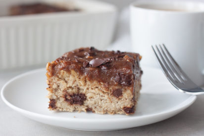 Slice of Date Caramel Chocolate Chip Baked Oatmeal on plate with fork, coffee in coffee cup, pan of oatmeal bake